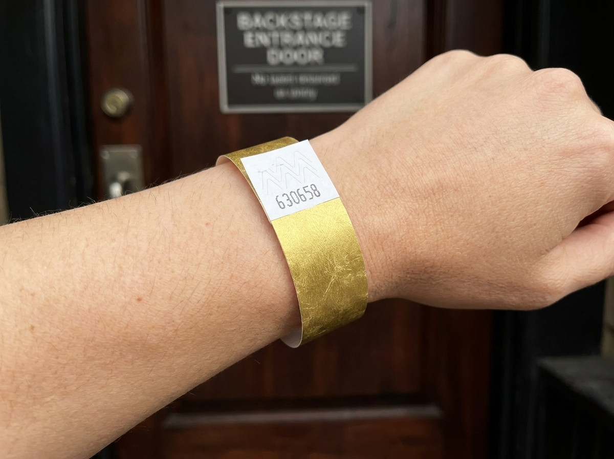 Pulsera VIP de festival en con impresión metalizada dorada y código de barras, llevada en una muñeca frente a una entrada backstage